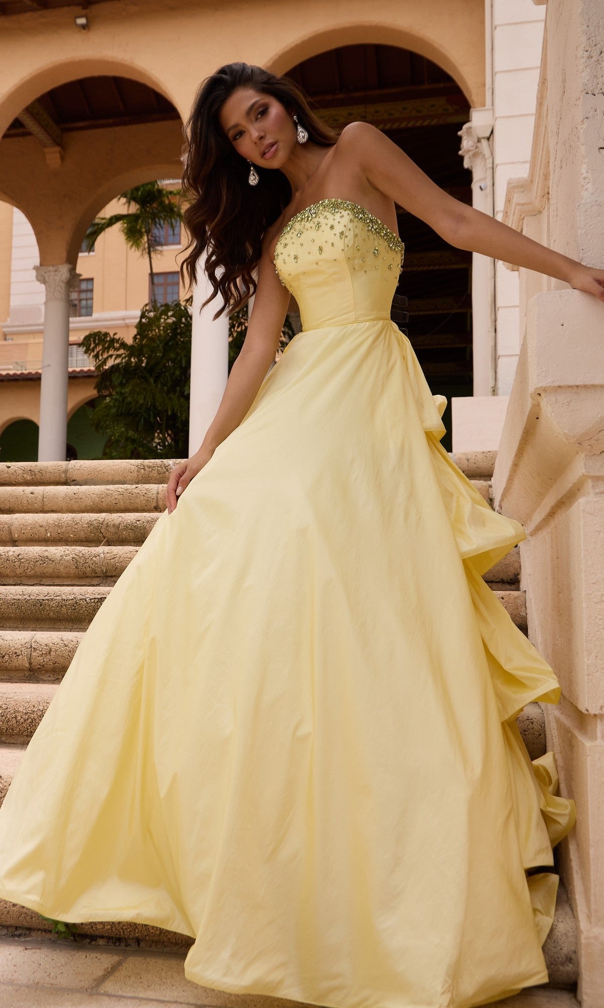 Woman in a Long Prom Dress (Ava Presley 26417) in soft yellow stands on stone steps, wearing a strapless ruffle ball gown with jeweled bodice, in front of an arched building.